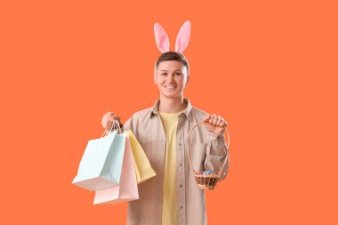 Tavşan kulaklı, alışveriş torbaları ve turuncu arka planda yumurtaları olan genç bir adam. Paskalya Satışı