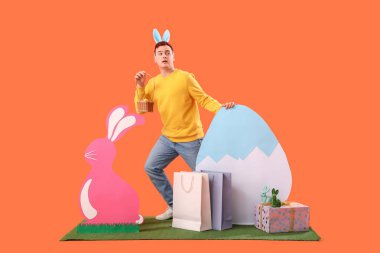 Tavşan kulaklı, sepetli ve çimlerin üzerinde turuncu arka planda dekoru olan genç bir adam. Paskalya Satışı