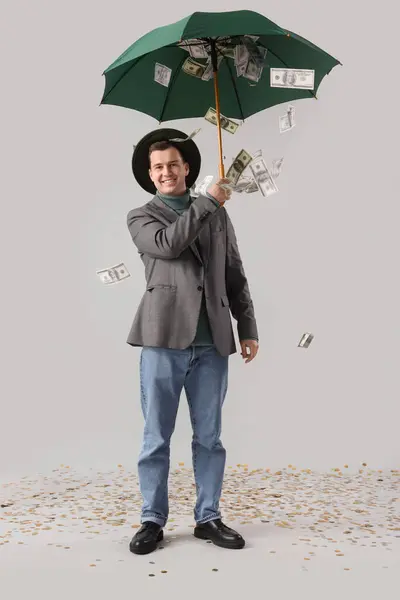 Young man with umbrella and falling money on light background