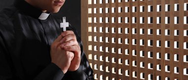 Young priest with cross in confession booth, closeup clipart