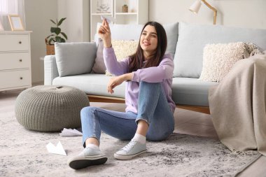 Evinde kağıt uçakla oturan genç bir kadın.