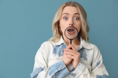 Şok olmuş genç bir kadın mavi arka planda büyüteçle, yakın plan.