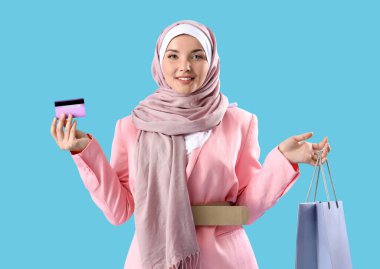 Mavi arka planda alışveriş çantası ve kredi kartıyla güzel bir Müslüman kadın.