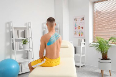 Fizyoterapi bandı olan genç bir adam rehabilitasyon merkezindeki kanepede oturuyor.