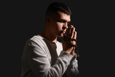 Tespihi olan genç adam karanlık arka planda dua ediyor.