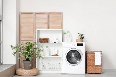 Sepetli modern çamaşır makinesi, raf ünitesi ve beyaz duvarın yanındaki giyinme ekranı. Ev çamaşır odasının içi.