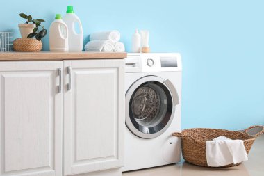 Mavi duvarın yanında sepeti ve dolabı olan modern çamaşır makinesi. Ev çamaşır odasının içi.