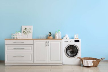 Mavi duvarın yanında sepeti ve dolapları olan modern çamaşır makinesi. Ev çamaşır odasının içi.