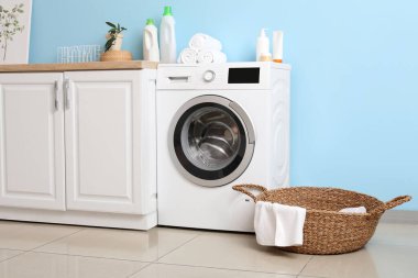 Mavi duvarın yanında sepeti ve dolabı olan modern çamaşır makinesi. Ev çamaşır odasının içi.
