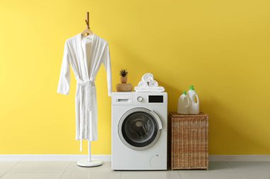 Çamaşır odasının içinde hasır sepeti, çamaşır makinesi ve bornoz var.