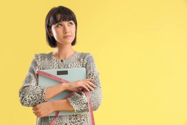 Ağırlık tartısı ve sarı arka planda mezura olan genç bir kadın. Diyet kavramı