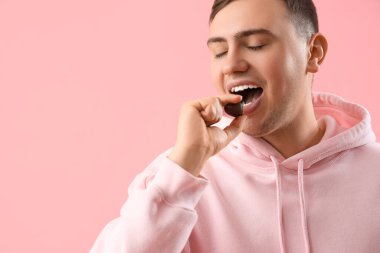 Genç adam pembe arka planda kalp şeklinde çikolata yiyor, yakın plan.