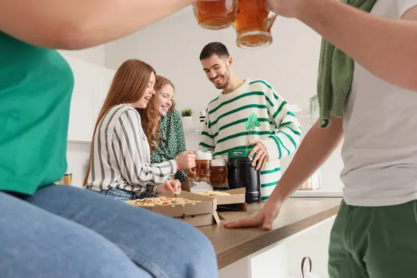 Group of young friends with beer and pizza celebrating St. Patrick\'s Day in kitchen
