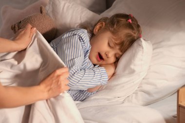 Anne, uyuyan kızını battaniyeyle örtüyor.