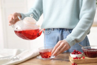 Genç bir kadın mutfakta çaydanlıktan fincana kızılcık çayı dolduruyor.