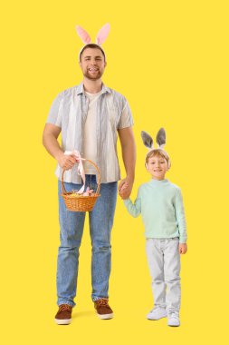 Mutlu baba ve küçük oğlu, sarı arka planda el ele tutuşan Paskalya yumurtalarıyla tavşan kulağı takıyor.