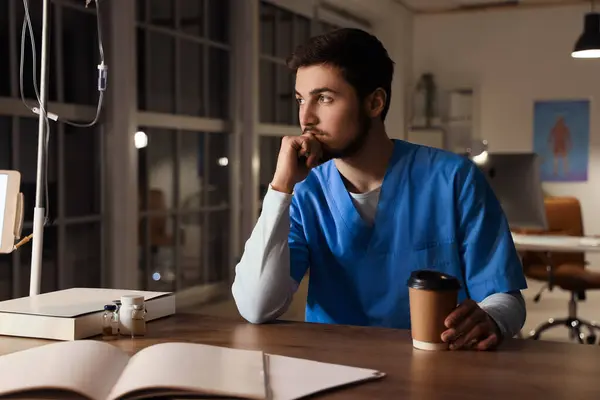 Male nurse with coffee cup working evening shift at desk in clinic