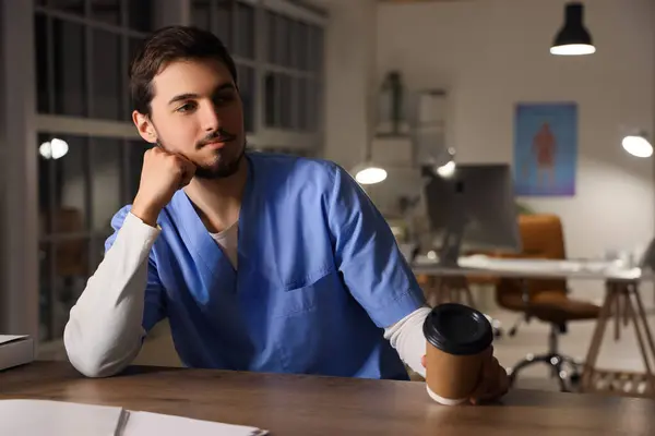Male nurse with coffee cup working evening shift at desk in clinic