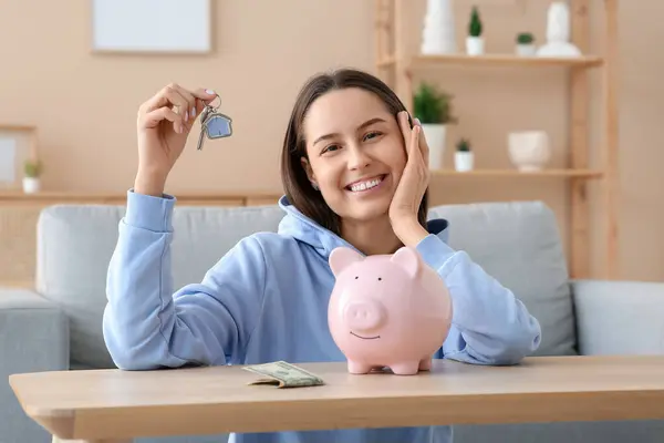 Pretty young woman with keys and piggy bank at home. Mortgage concept