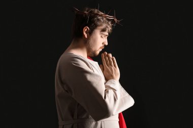 İsa cüppeli ve dikenli taç giyen adam kara arka planda ışıkla dua ediyor.