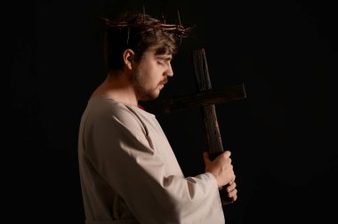 İsa cüppeli bir adam ve dikenli bir taç, siyah arka planda ışıkla ahşap bir haç taşıyor.