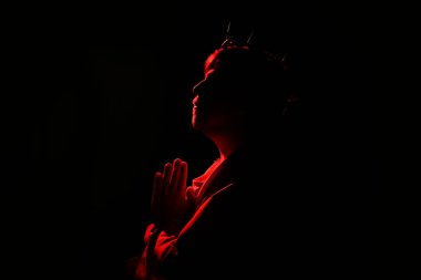 İsa cüppeli ve dikenli taç giyen adam siyah arka planda kırmızı ışıkla dua ediyor.