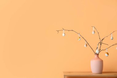 Ağaç dalları ve bej duvarın yanındaki rafta Paskalya yumurtaları olan vazo.