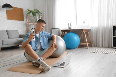 Evde dizüstü bilgisayarı, su şişesi ve fitbol topu olan yakışıklı bir adam.