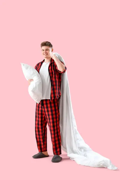 Young man in pajamas with soft pillow and blanket on pink background