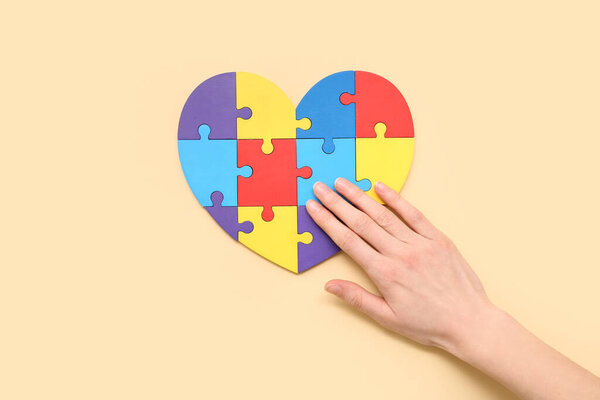 Female hand touching heart made of color puzzle on light yellow background. Concept of autistic disorder
