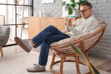 Orta yaşlı bir adam bir bardak suyla oturma odasında haplara bakıyor.