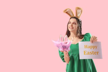 Tavşan kulaklı güzel bir kadın elinde kartla Mutlu Paskalyalar ve pembe arka planda pasta.