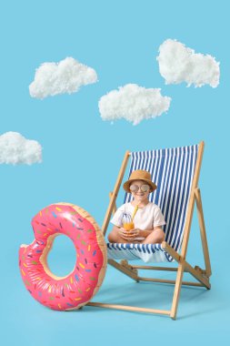 Kokteyl ve yüzme yüzüğüyle şirin bir çocuk mavi arka planda iskemlede oturuyor.