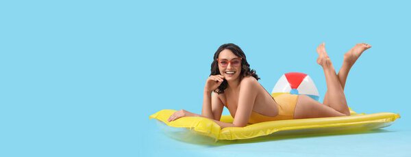 Pretty young woman in swimsuit lying on inflatable mattress against light blue background with space for text