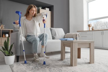 Koltuk değnekleriyle yaralanmış genç bir kadın. Evdeki koltukta oturuyor.