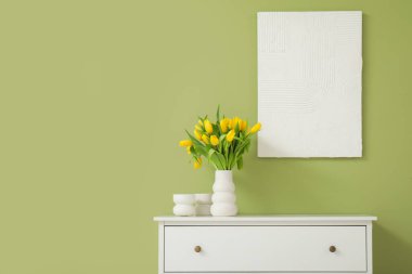 Çekmece dolabında sarı lale vazosu ve oturma odasındaki duvarda resim.