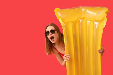 Tank mayosu giymiş, kırmızı arka planda şişme yatağı olan güzel, şaşkın genç bir kadın.