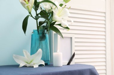 Boş cenaze çerçevesi, yanan mum ve vazo. Renk duvarının yanındaki masada zambak çiçekleri var.