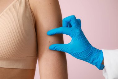 Dermatolog pembe arka planda benleri olan genç bir kadını muayene ediyor.