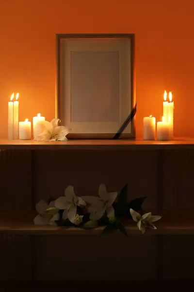Blank funeral frame, burning candles and lily flowers on wooden table in dark room