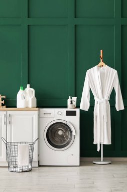 Çamaşır sepeti, çamaşır makinesi ve bornozlu odanın içi.