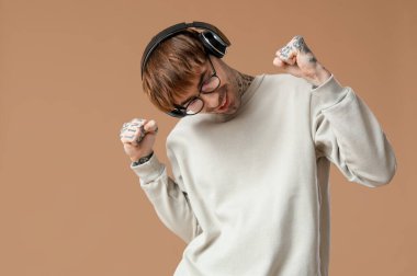 Kulaklıklı genç dövmeli adam bej arka planda dans ediyor.