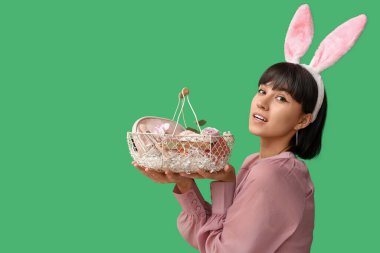 Tavşan kulaklı güzel bir kadın yeşil arka planda kozmetik ürünleriyle Paskalya sepeti tutuyor.