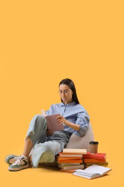 Gözlüklü genç kadın sarı arka planda kitap okuyor.