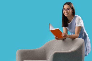 Gözlüklü genç kadın mavi arka planda koltuğun arkasında kitap okuyor.