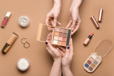 Farklı dekoratif kozmetik ürünleri ve kahverengi arka planda küpeleri olan kadın elleri.