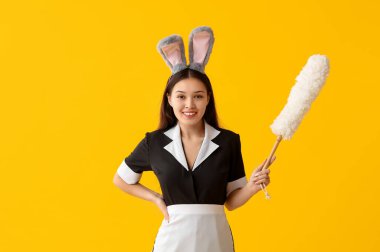 Tavşan kulaklı genç Asyalı oda hizmetçisi ve sarı arka planda toz bezi. Paskalya kutlaması