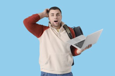 Mavi arka planda dizüstü bilgisayarı ve sırt çantası olan kafası karışmış bir erkek turist.