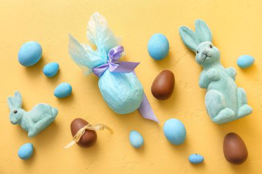 Kağıda sarılmış boyanmış Paskalya yumurtaları ve sarı arka planda oyuncak tavşancıklar.