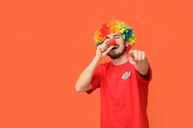 Komik palyaço kılığına girmiş yakışıklı genç adam turuncu arka plandaki izleyiciyi işaret ediyor. 1 Nisan kutlamaları